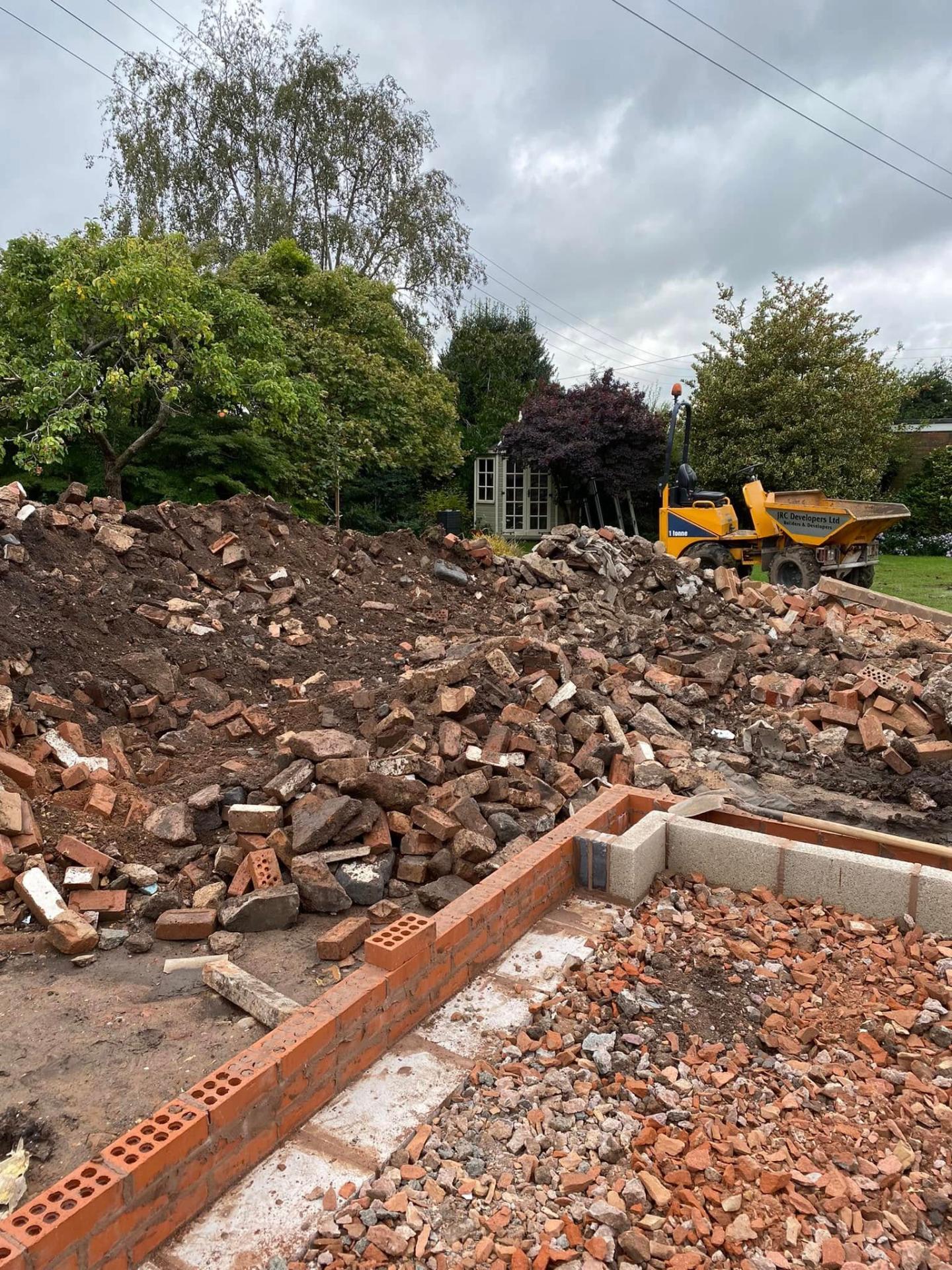 Large pile of demolition brick and rubble at White Gates with a yellow JRC Developers Ltd mini-dumper working in the background against new red brick footings