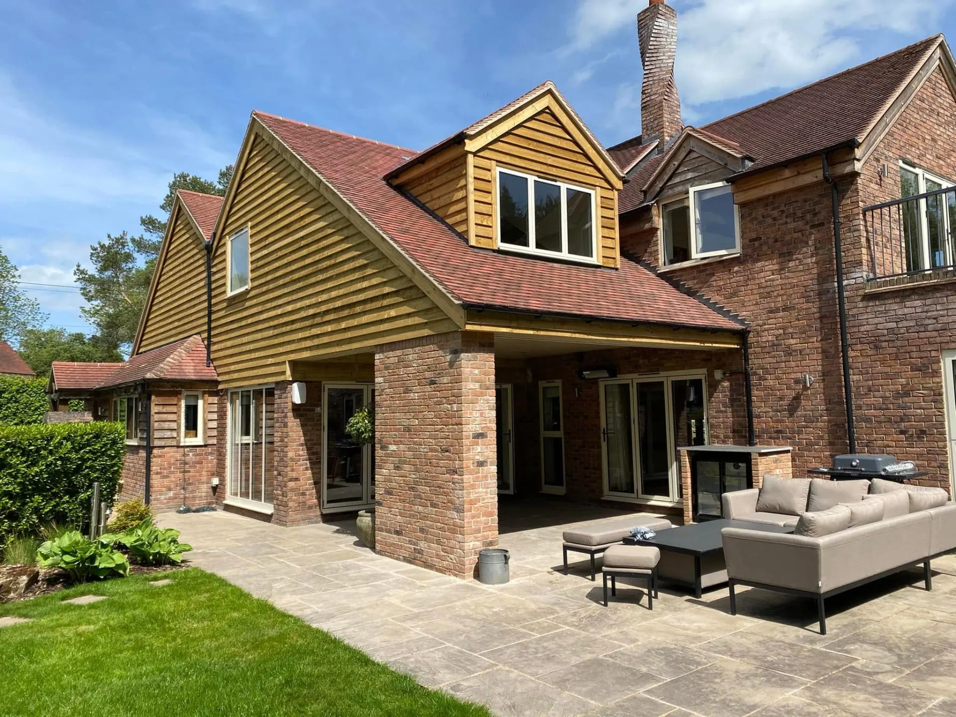 Rear elevation of Dingleside in Worcestershire showing a new two-bedroom extension with oak-clad gable and dormer, plain clay tile roof, brick columns and a covered outdoor entertainment terrace with bifold doors — built by JRC Developers