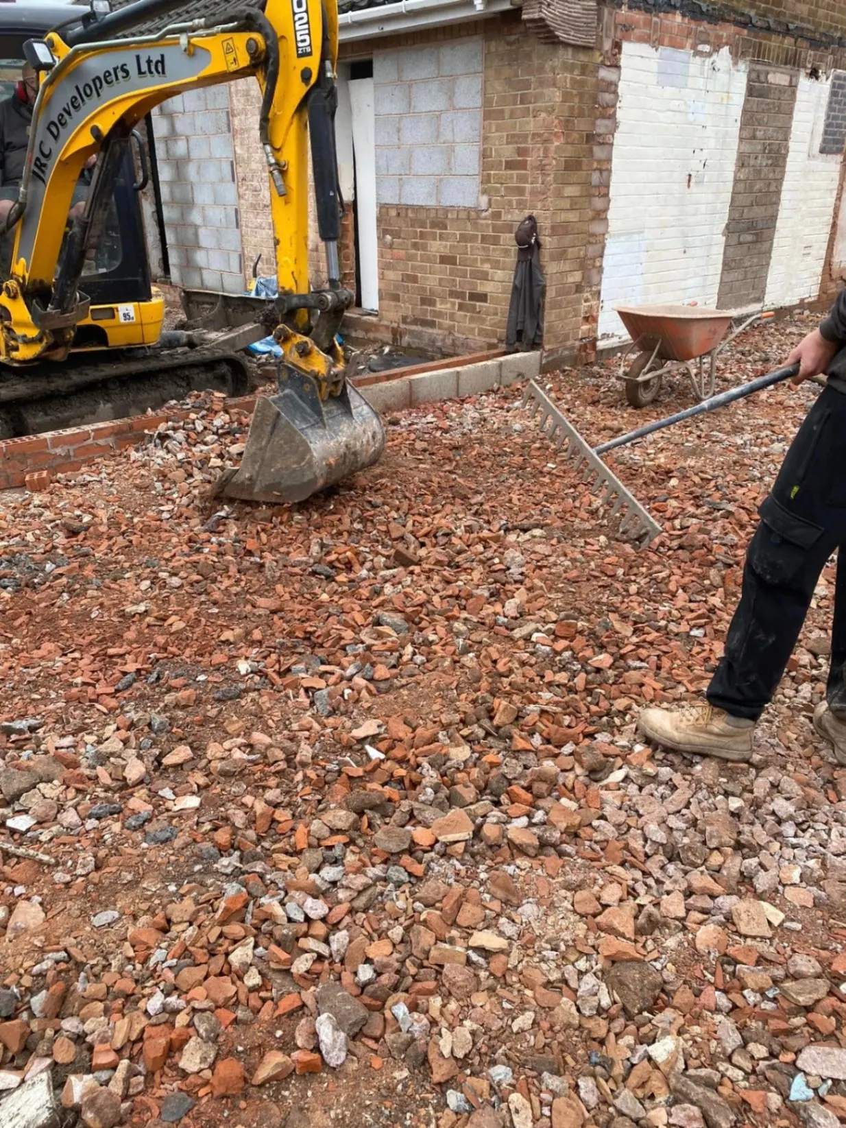 JRC Developers mini-excavator raking crushed brick sub-base at White Gates alongside a worker with a landscaping rake, in front of the stripped bungalow shell