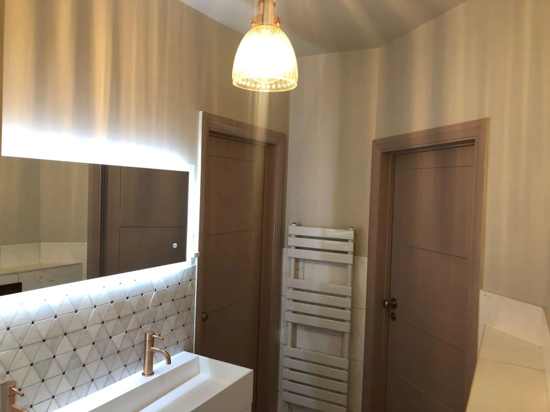Bathroom detail showing illuminated mirror, feature tiled splashback, brass tapware and timber-toned doors