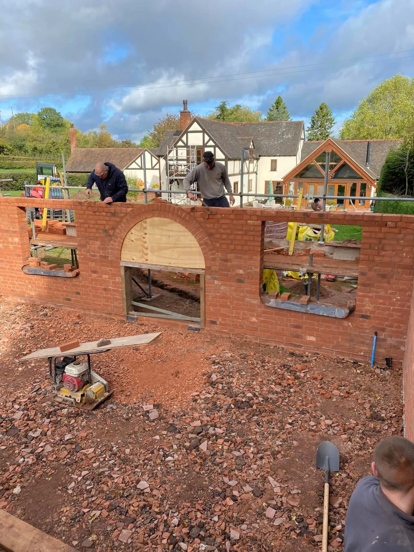 Two bricklayers laying the man cave walls with a turned brick arch over the doorway and a black and white timbered house behind
