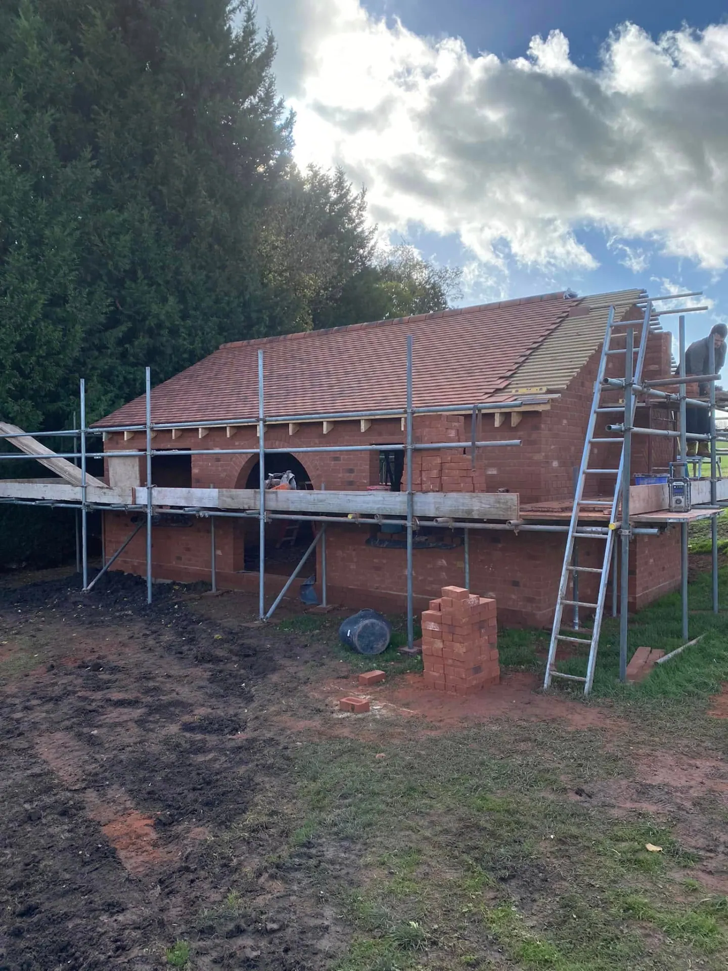 Roofers tiling the man cave with clay tiles on battens, scaffold and ladder in place around the red brick outbuilding