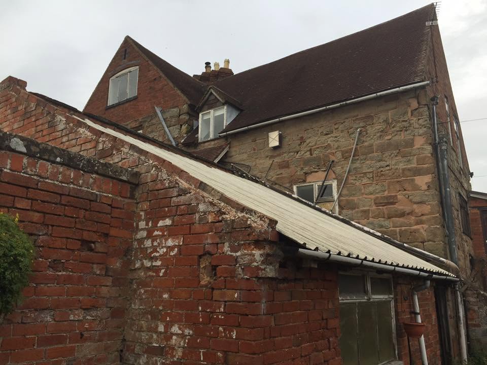 Corrugated asbestos-cement lean-to roof running against the historic brick boundary wall of the Grade II listed farmhouse before removal