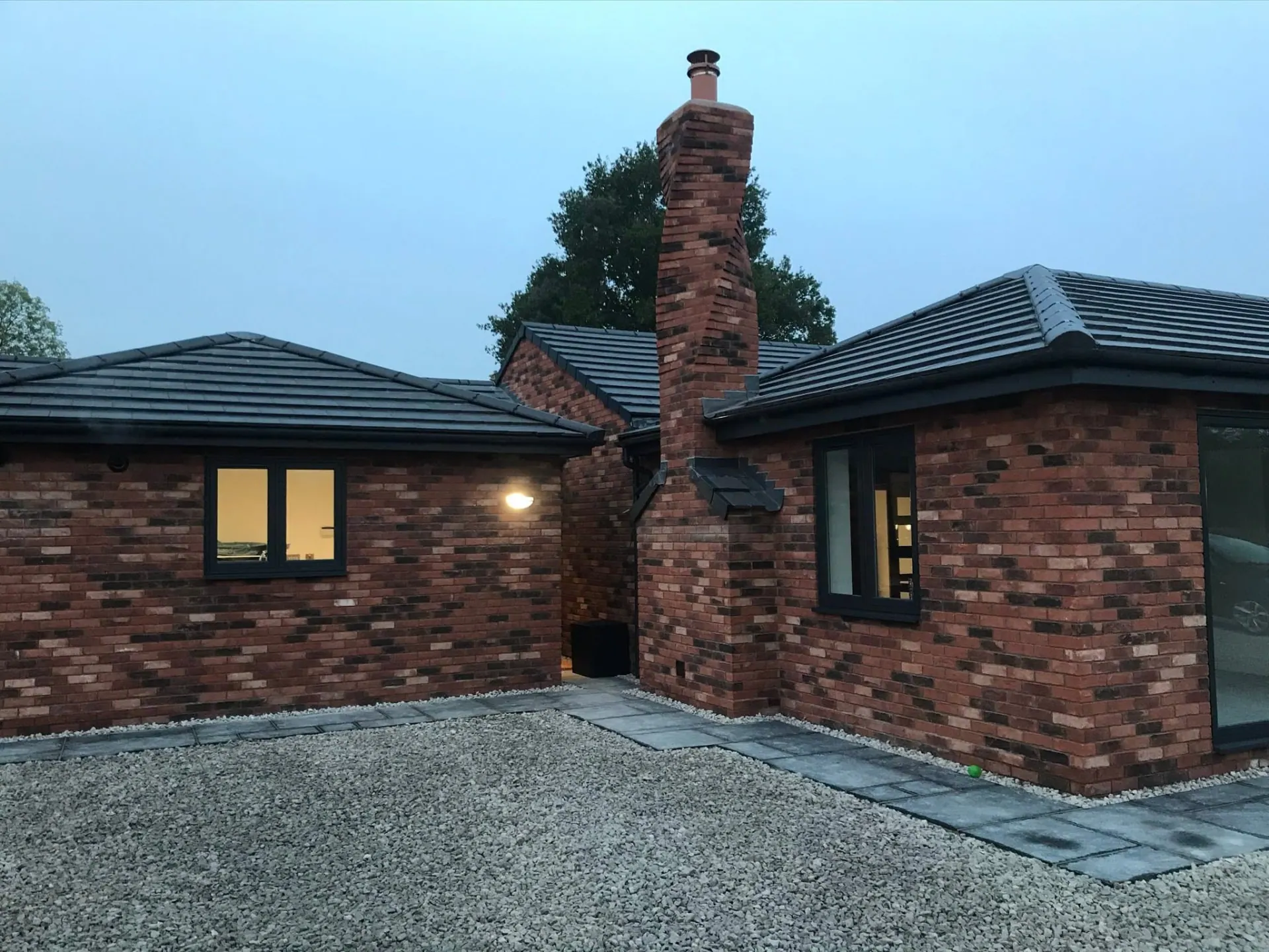 Brick bungalow at dusk showing the completed hand-laid twisted brick chimney stack, lead flashings and gravel courtyard