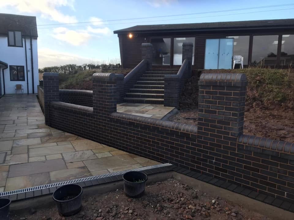 Blue brick boundary and retaining walls with capped pillars, steps and a slot drainage channel set into an Indian sandstone patio