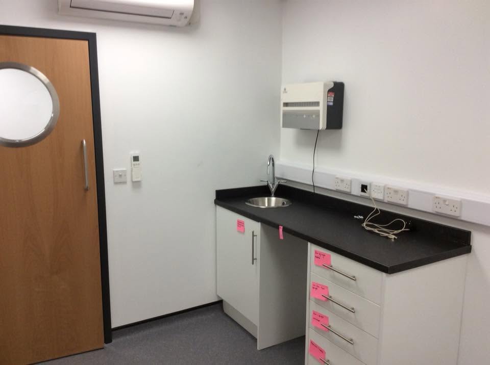Consult room fitted with dark worktop, stainless sink, white base units and wall-mounted air conditioning unit