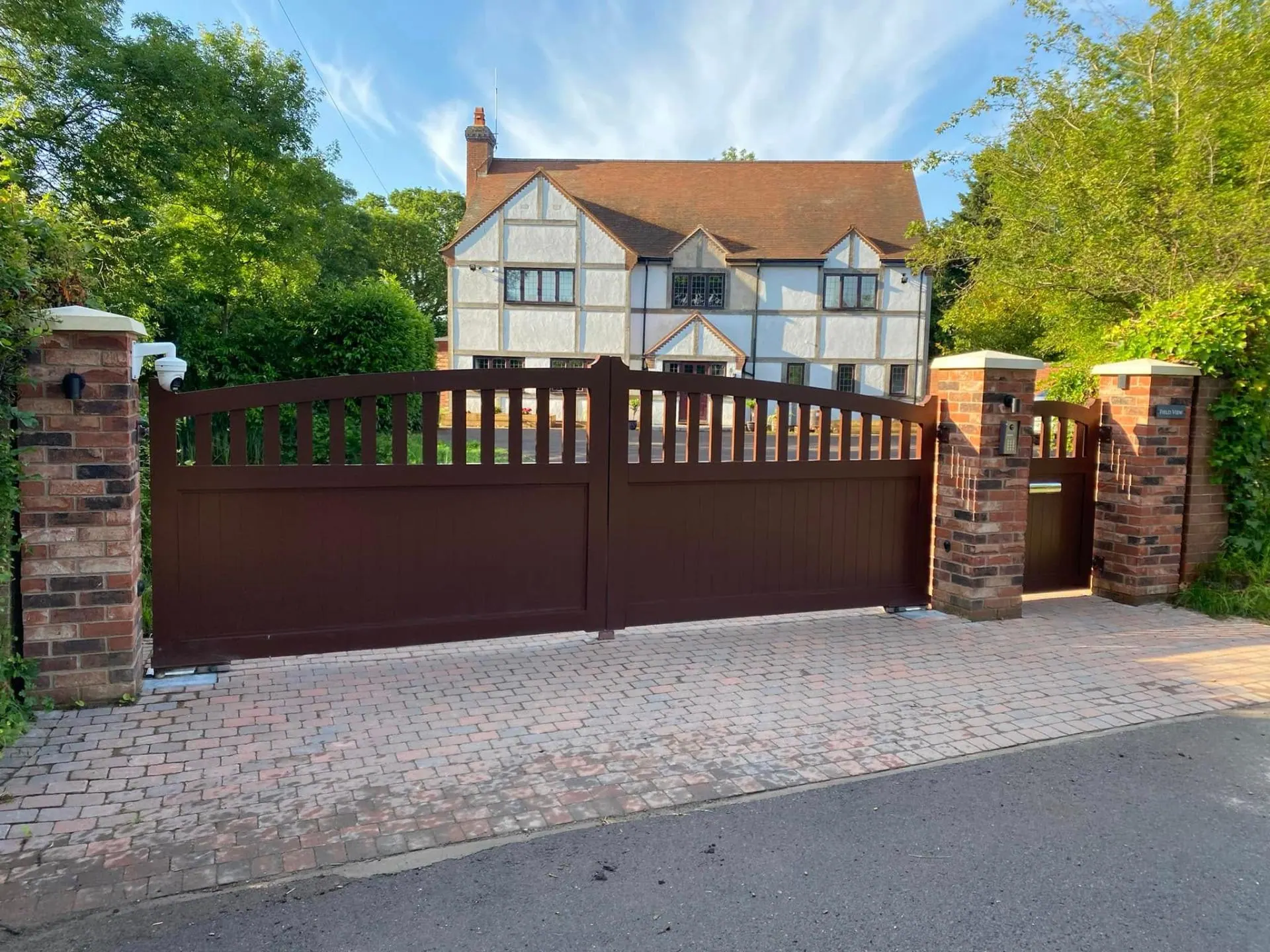 Automated timber driveway gates in dark oxblood finish with brick pillars, intercom and CCTV outside Field View — a Tudor-style home in Worcestershire built by JRC Developers
