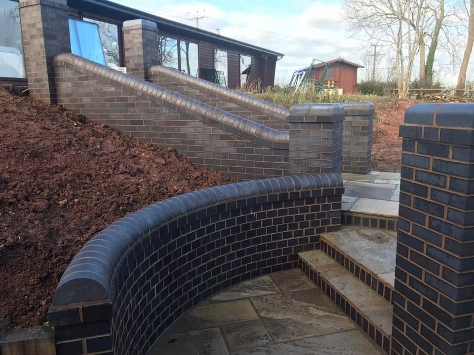 Curved blue engineering brick retaining walls with bullnose coping, brick and Indian sandstone garden steps and a riven sandstone terrace built by JRC Developers in Worcestershire