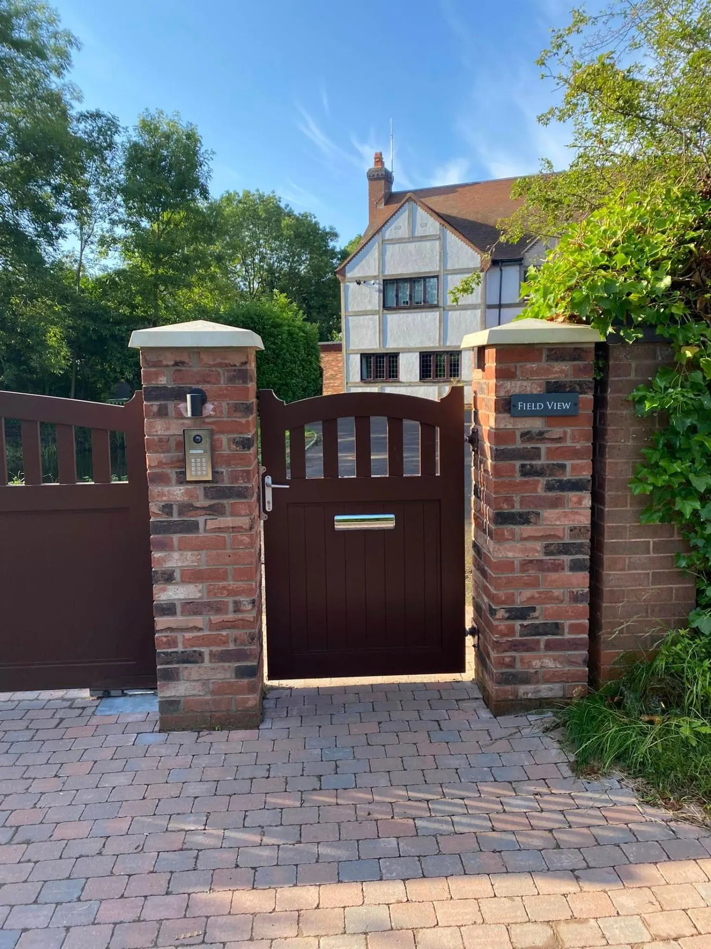 Close-up of the pedestrian gate at Field View, showing the arched top rail, dark oxblood timber, stainless letterplate, brass Field View name plaque and pillar-mounted video intercom with keypad