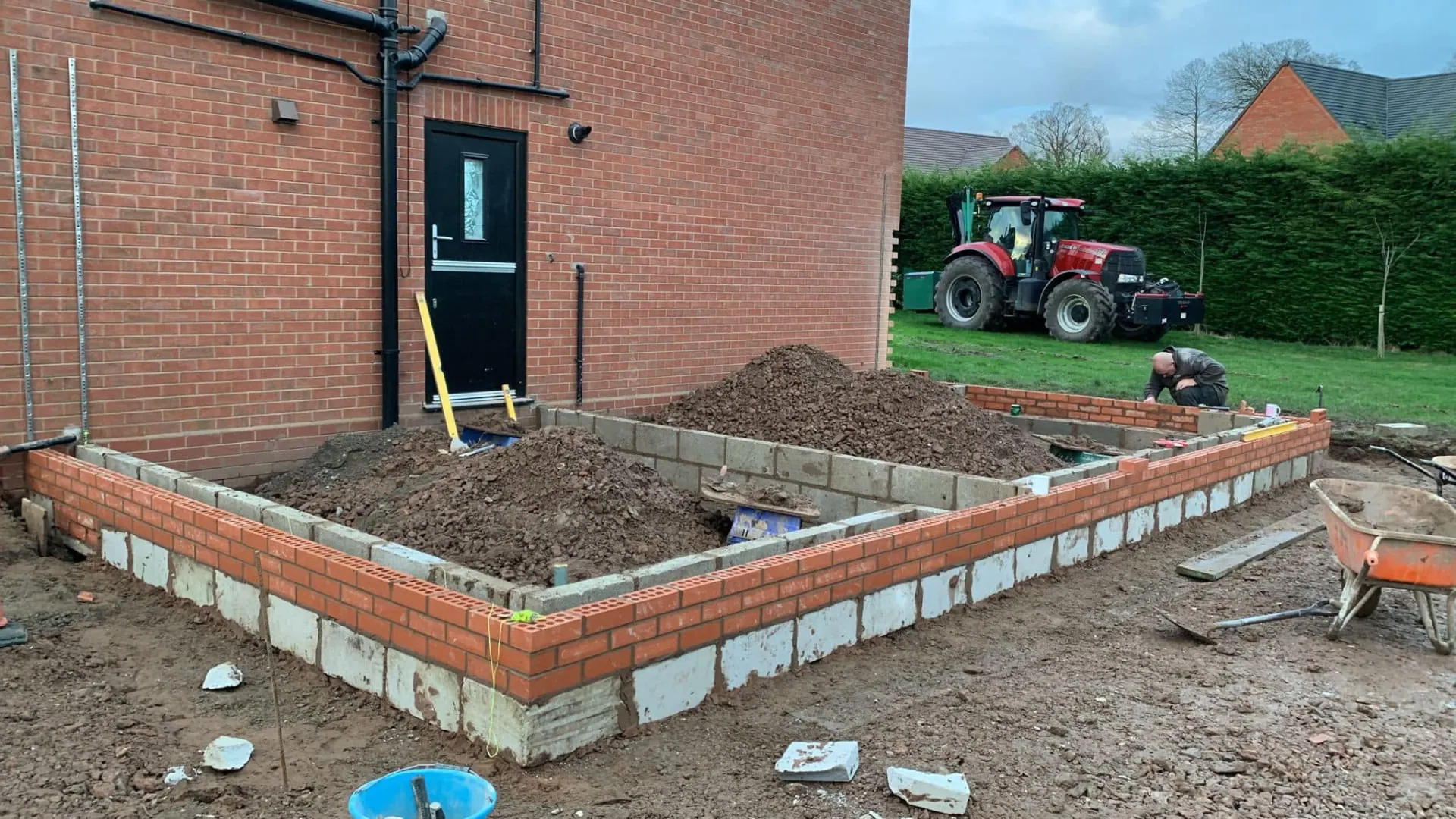 Brick and block foundation walls built off concrete footings for a side extension, with drainage and a builder working in the background