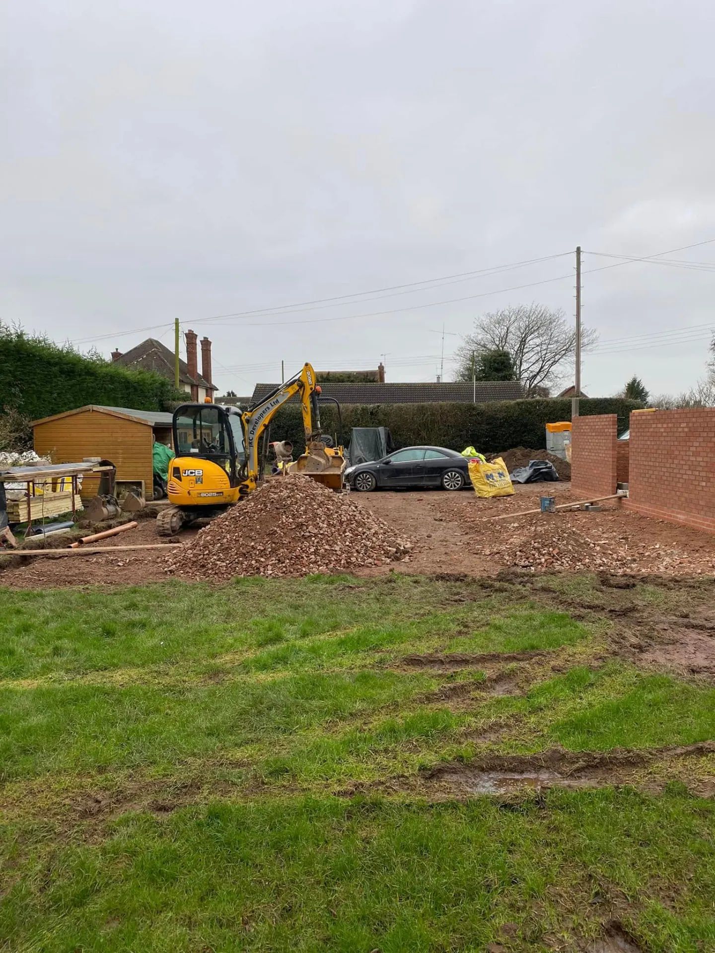 Wide view of the working site showing the excavator, stone stockpile and the new red brick boundary wall under construction