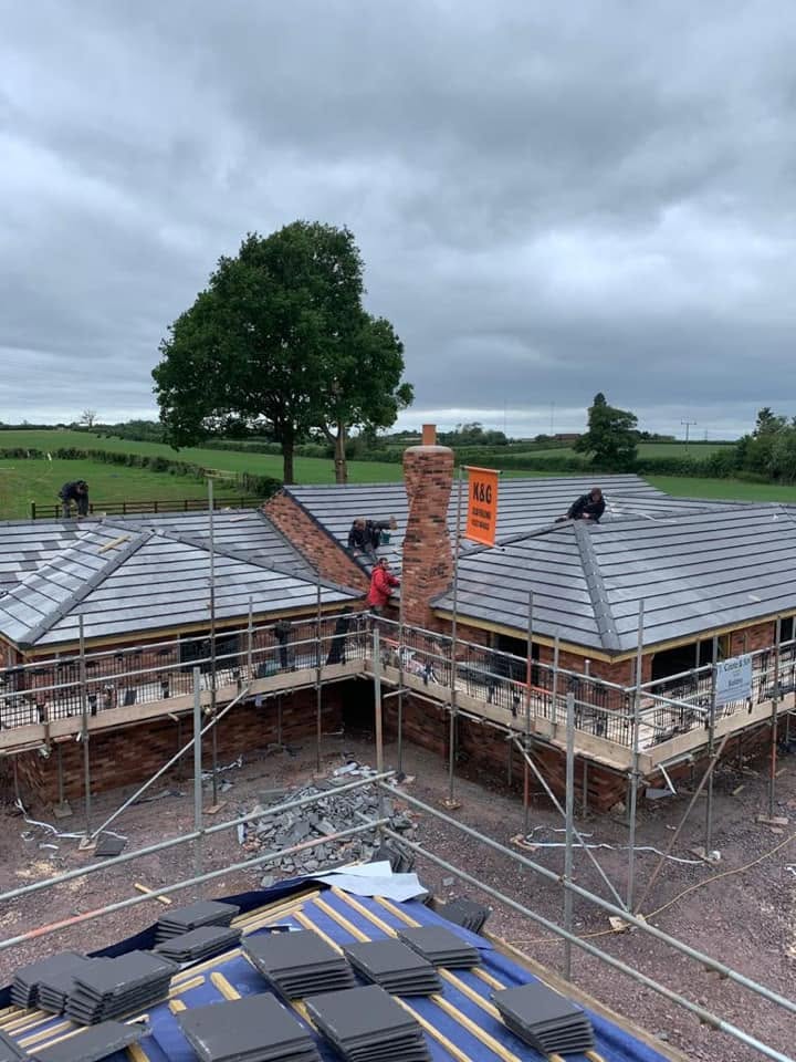 Roofing crew tiling the hipped roofs of a new-build brick bungalow from scaffolding, with the brick chimney stack rising through the roof line