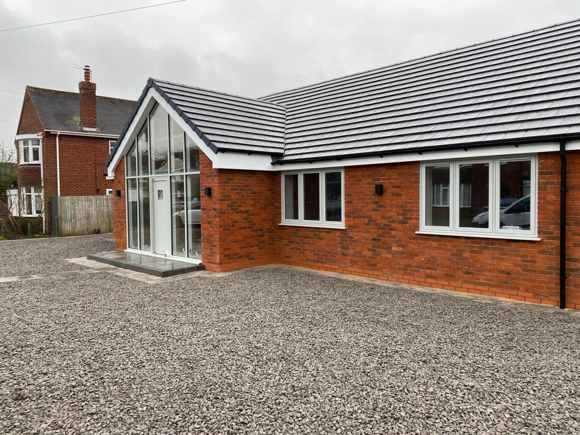 Opposite-side view of the finished White Gates bungalow showing the glazed gable entrance, new grey plain tile roof, red brick corner detailing and gravel drive extending to the neighbours' boundary